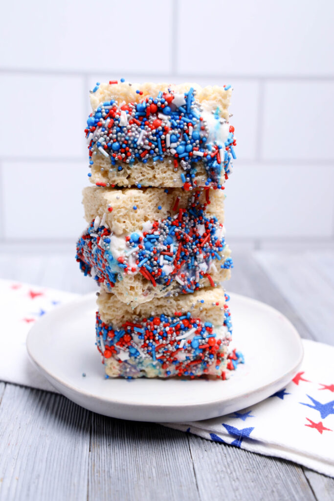 July 4th Rice Krispies Ice Cream Sandwiches 8 Three stacked No-Bake Patriotic Rice Krispies treats covered in red, white, and blue sprinkles on a white plate, with a star-patterned cloth and white tile wall in the background.
