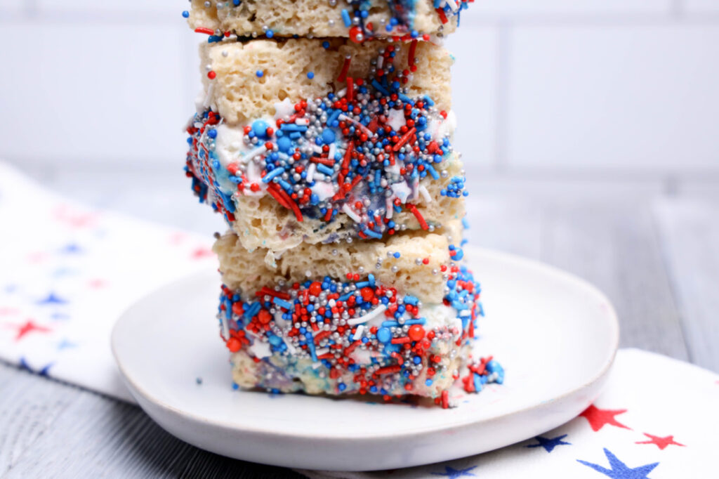 July 4th Rice Krispies Ice Cream Sandwiches 9 Three No-Bake Patriotic Rice Krispies treats stacked on a white plate, covered in white icing and red, white, and blue sprinkles.