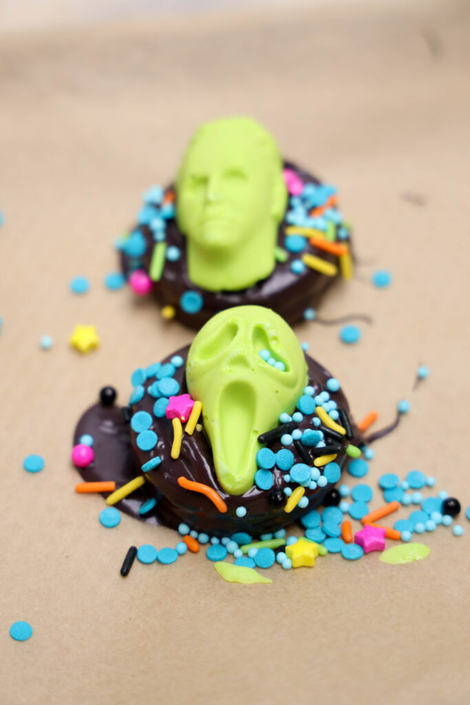 Summerween Slasher Oreos 7 Two Summerween Slasher Oreos topped with green candy faces, colorful sprinkles, and star-shaped decorations rest on a brown surface.