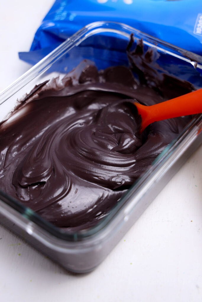 Summerween Slasher Oreos 5 A glass dish filled with smooth, melted dark chocolate being stirred with an orange spatula—perfect for dipping Summerween Slasher Oreos or your favorite spooky Summerween treats.