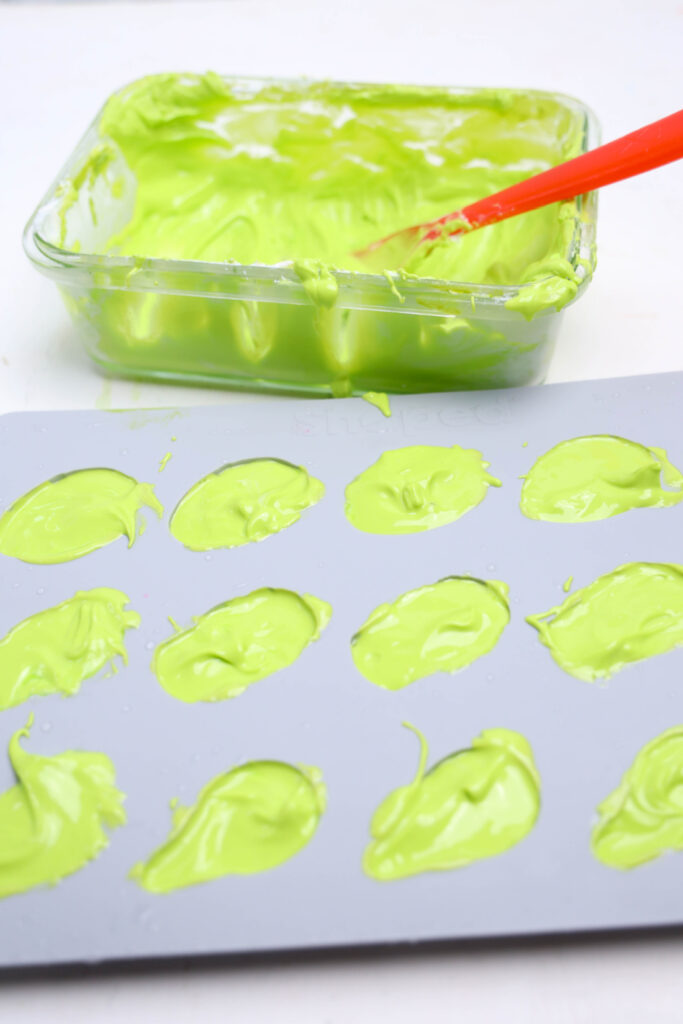 Summerween Slasher Oreos 4 A silicone mold filled with green creamy mixture sits in front of a glass dish containing more of the mixture, a red spatula, and crushed Oreos—perfect for your next Summerween treat.