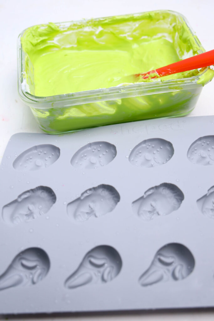 Summerween Slasher Oreos 3 A gray silicone mold with face-shaped cavities sits next to a glass dish of green mixture and a red spatula, ready to craft spooky Summerween Slasher Oreos.