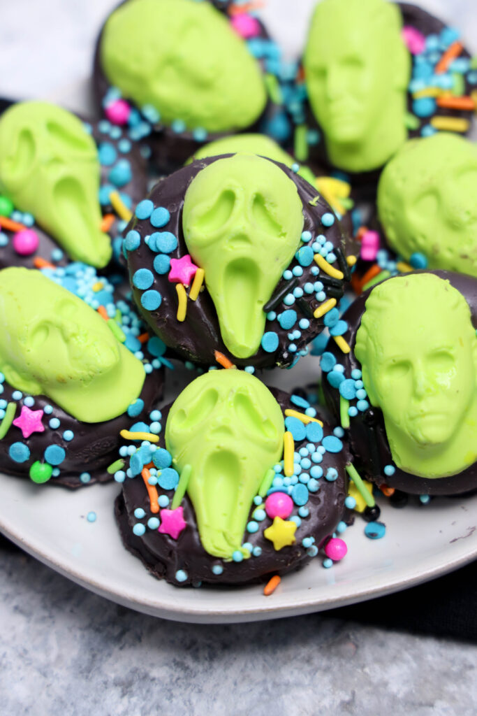 Summerween Slasher Oreos 8 Chocolate-covered cookies, called Summerween Slasher Oreos, are topped with scream-faced and skull-shaped candies, plus colorful sprinkles, served on a white plate—perfect for a spooky Summerween treat!.