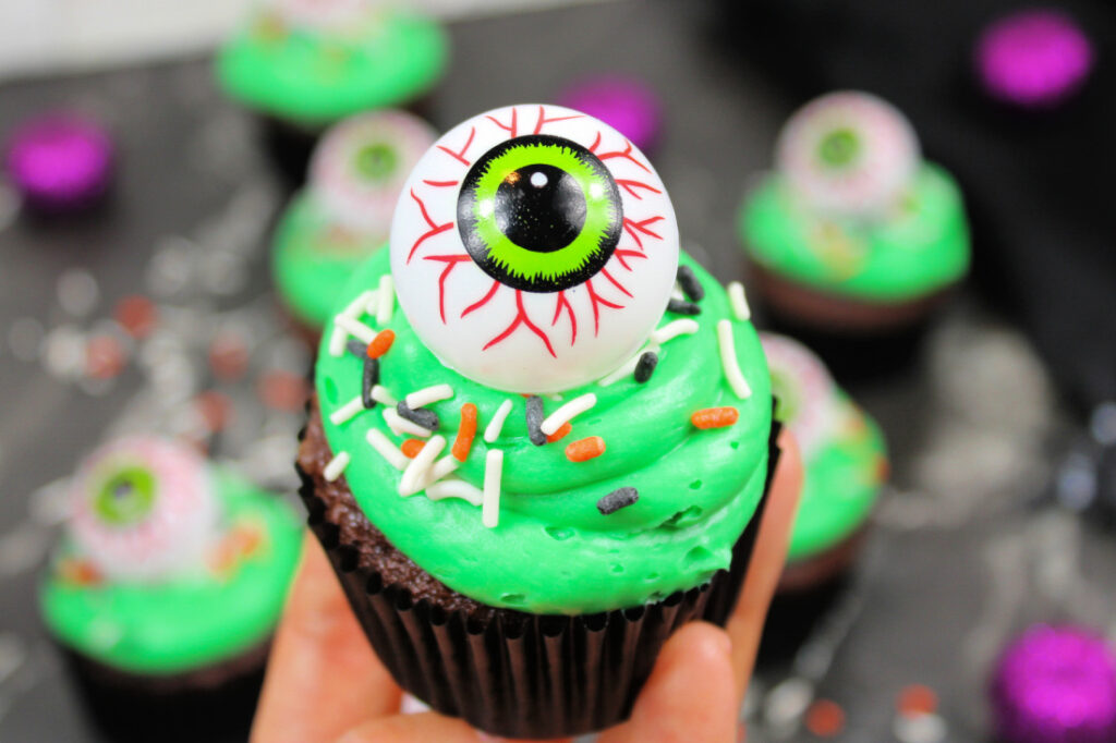 Spooky Halloween Eyeball Cupcakes: The Perfect Creepy-Cute Treats 2 A chocolate cupcake with green frosting, colorful sprinkles, and a large plastic eyeball decoration on top, held in a person's hand—perfect for fans of Spooky Halloween Eyeball Cupcakes.