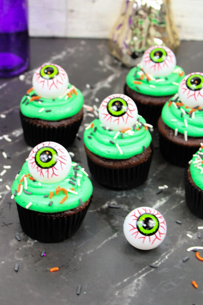 Spooky Halloween Eyeball Cupcakes: The Perfect Creepy-Cute Treats 6 Spooky Halloween Eyeball Cupcakes with green frosting, topped with plastic eyeball decorations and colorful sprinkles, are arranged on a dark surface.