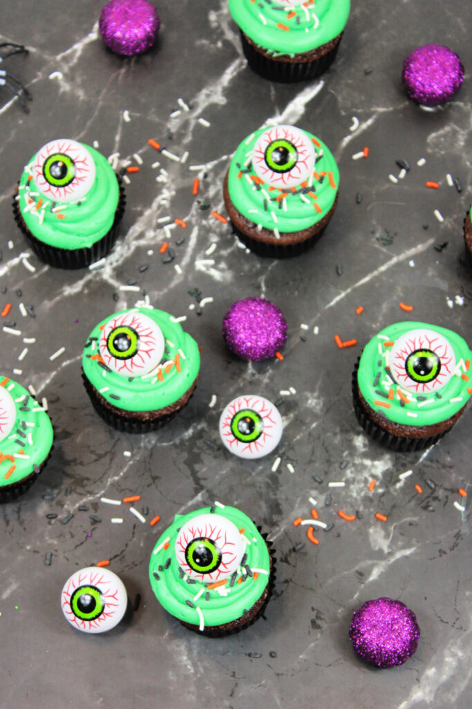 Spooky Halloween Eyeball Cupcakes: The Perfect Creepy-Cute Treats 7 Spooky Halloween Eyeball Cupcakes with green frosting, candy eyeball toppers, Halloween-themed sprinkles, and purple glittery decorations are displayed on a dark marble surface.