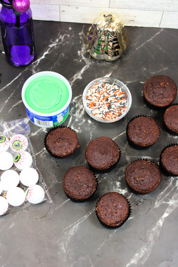 Spooky Halloween Eyeball Cupcakes: The Perfect Creepy-Cute Treats 3 Chocolate cupcakes on a dark counter with green frosting, Halloween sprinkles, candy eyeballs, a purple bottle, and a decorative item—perfect for making Spooky Halloween Eyeball Cupcakes that are sure to delight at any Halloween party.