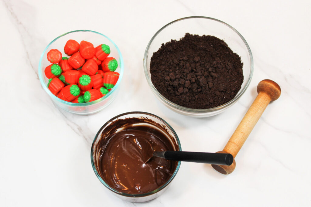 Easy Pumpkin Patch Brownies Recipe - Mini Halloween Treats (OMG So Cute!) 3 A bowl of pumpkin-shaped candies, a bowl of crushed chocolate cookies, and a bowl of melted chocolate with a spoon create the perfect setup for making festive mini Halloween brownies or fun pumpkin patch brownies on a white surface.
