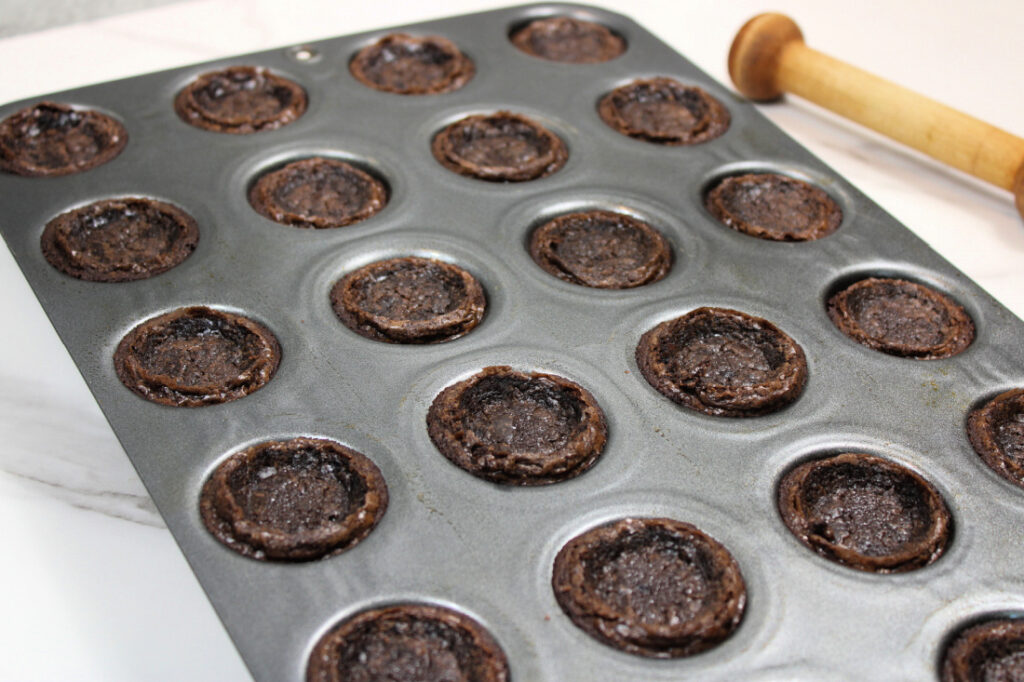 Easy Pumpkin Patch Brownies Recipe - Mini Halloween Treats (OMG So Cute!) 5 A mini muffin tin filled with partially baked chocolate cookie cups, perfect for creating mini halloween brownies, sits next to a wooden tool on a marble countertop.