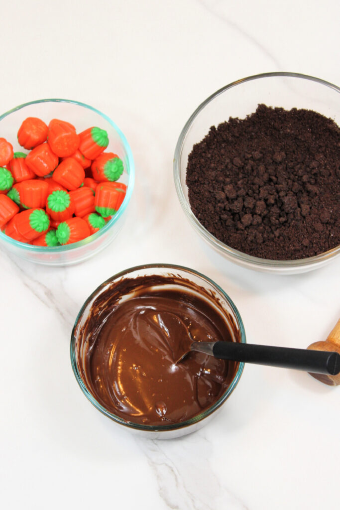 Easy Pumpkin Patch Brownies Recipe - Mini Halloween Treats (OMG So Cute!) 6 Three glass bowls on a white surface: one with pumpkin-shaped candies, one with crushed chocolate cookies, and one with melted chocolate—perfect for assembling festive mini Halloween brownies or creating your own pumpkin patch brownies.