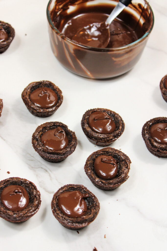 Easy Pumpkin Patch Brownies Recipe - Mini Halloween Treats (OMG So Cute!) 7 Chocolate-filled cookie cups are arranged on a white surface, resembling mini Halloween brownies, with a glass bowl of melted chocolate and a spoon in the background.