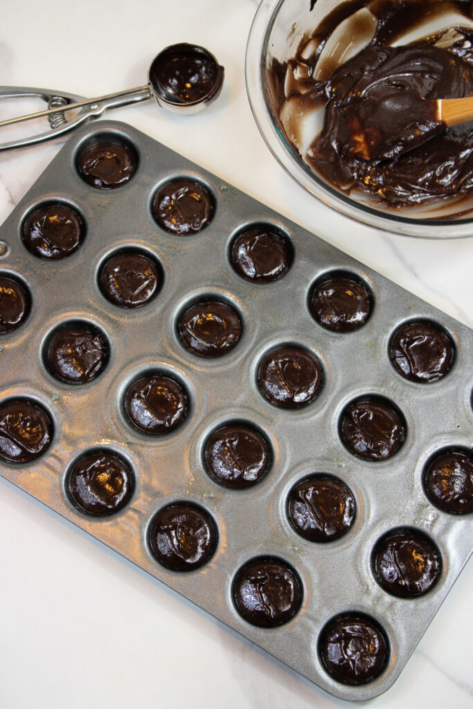 Easy Pumpkin Patch Brownies Recipe - Mini Halloween Treats (OMG So Cute!) 4 A muffin tin filled with chocolate batter for mini halloween brownies sits next to a bowl of leftover batter and a metal scoop on a white surface.
