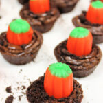 Easy Pumpkin Patch Brownies Recipe - Mini Halloween Treats (OMG So Cute!) 15 Chocolate brownie bites topped with orange and green pumpkin-shaped candies, arranged on a white surface with cookie crumbs, make these look just like pumpkin patch brownies—perfect mini Halloween brownies for your spooky celebration!.