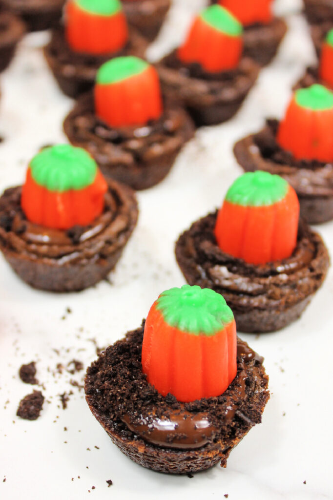 Easy Pumpkin Patch Brownies Recipe - Mini Halloween Treats (OMG So Cute!) 9 Chocolate brownie bites topped with orange and green pumpkin-shaped candies, arranged on a white surface with cookie crumbs, make these look just like pumpkin patch brownies—perfect mini Halloween brownies for your spooky celebration!.