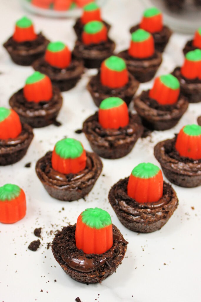 Easy Pumpkin Patch Brownies Recipe - Mini Halloween Treats (OMG So Cute!) 14 Mini Halloween brownies topped with orange pumpkin-shaped candies, arranged on a white surface with scattered brownie crumbs, create a festive pumpkin patch brownies display.