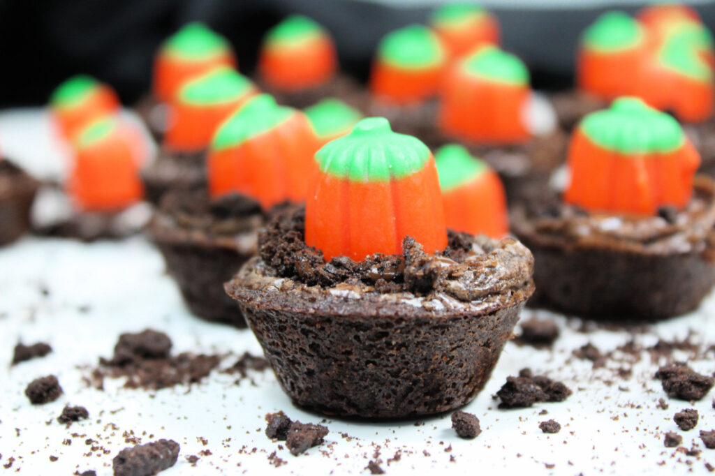 Easy Pumpkin Patch Brownies Recipe - Mini Halloween Treats (OMG So Cute!) 2 Mini Halloween brownies are topped with crushed cookies and candy pumpkins, creating adorable pumpkin patch brownies arranged on a white surface with cookie crumbs scattered around.