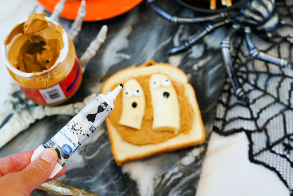 A slice of Halloween Toast with peanut butter is topped with two banana ghosts decorated with icing. In the foreground, a hand holds a tube of black decorating gel, perfect for spooky finishing touches.