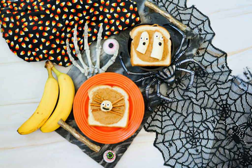 Two slices of Halloween Toast decorated as spooky creatures rest on a black surface, surrounded by bananas, skeleton hands, fake eyeballs, and spider web decorations.