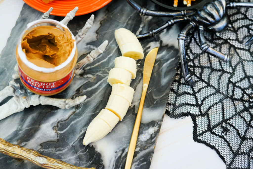 A sliced banana on a marble surface with a gold knife, an open jar of peanut butter, plastic skeleton hands, a fake spider, and black lace web decor—perfect for crafting spooky Halloween Toast.