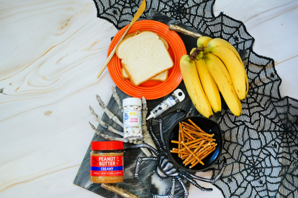 A Halloween Toast setup featuring bread, peanut butter, bananas, candy eyes, pretzel sticks, and skeleton hand props arranged on a web-patterned cloth for a spooky snack display.