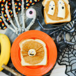 Two slices of Halloween Toast with peanut butter are decorated as festive snacks: one as a spider with pretzel legs, the other as ghosts made from banana slices and chocolate chips for spooky faces.