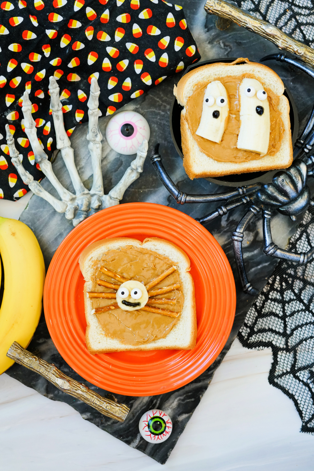Two slices of Halloween Toast with peanut butter are decorated as festive snacks: one as a spider with pretzel legs, the other as ghosts made from banana slices and chocolate chips for spooky faces.