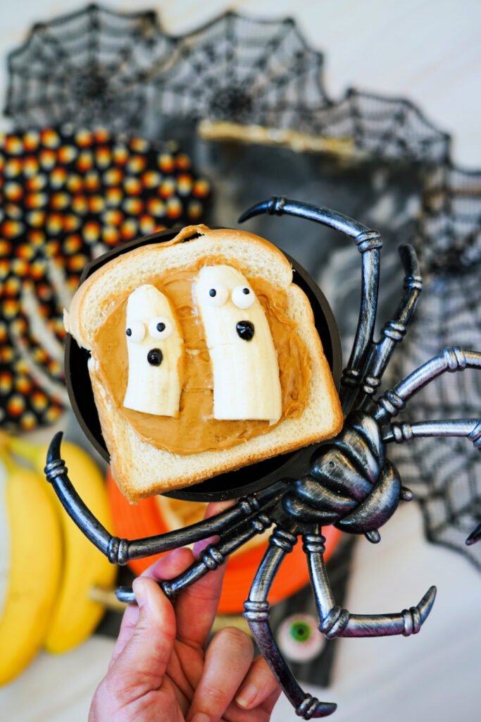 A hand holds a slice of Halloween Toast with peanut butter and two banana slices decorated as ghosts. A large fake spider and spooky, Halloween-themed decorations fill the background.