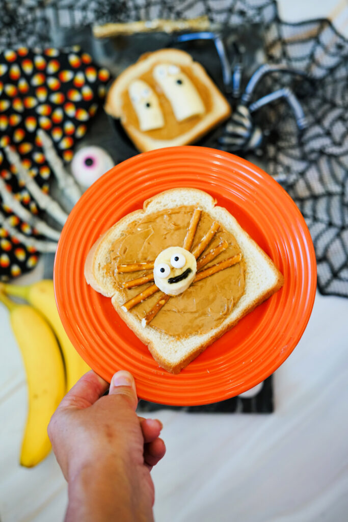 A hand holds an orange plate with a slice of Halloween Toast—bread topped with peanut butter, pretzel sticks, and a candy eye arranged like a spider. Halloween-themed food items are visible in the background.