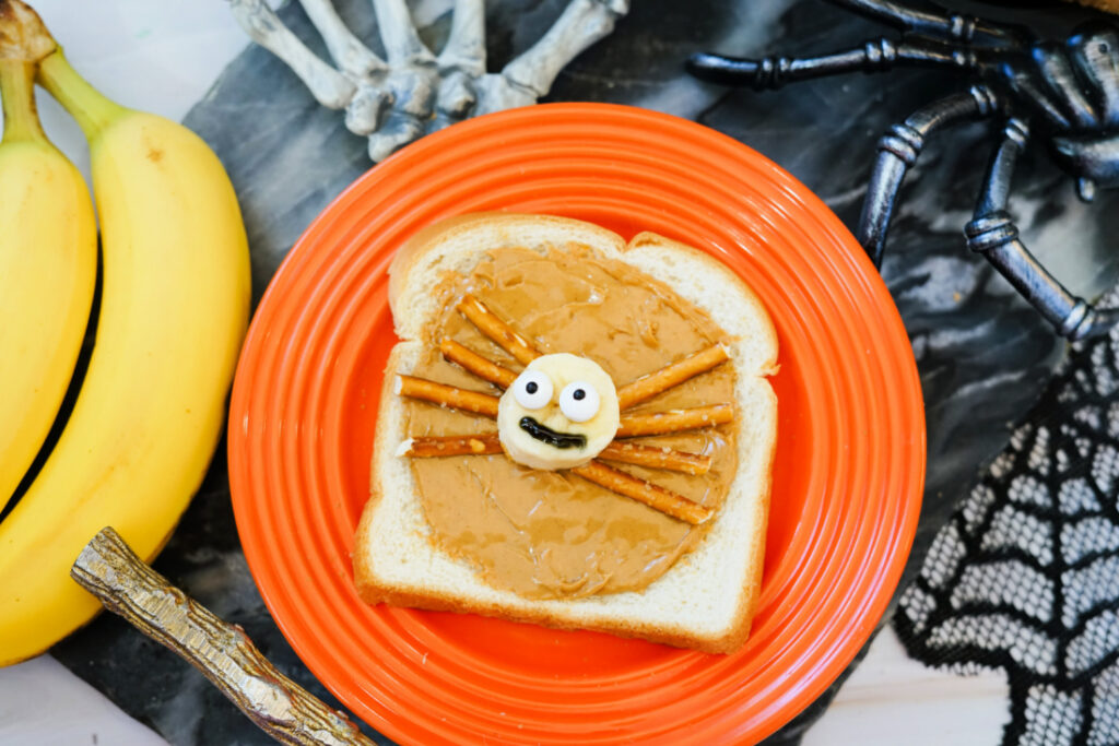 A slice of Halloween Toast with peanut butter, topped with banana slices, pretzel stick legs, and candy eyes to resemble a spider, served on an orange plate surrounded by bananas and festive Halloween decorations.
