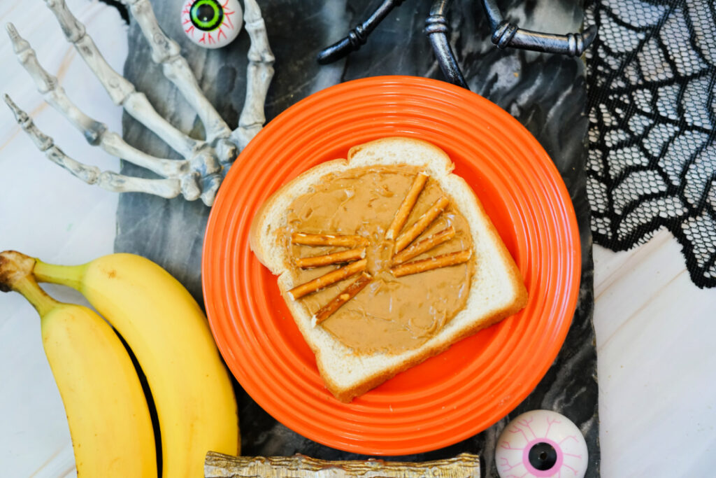 A festive Halloween Toast: a slice of bread with peanut butter and pretzel sticks arranged like a spider on an orange plate, surrounded by bananas, fake eyeballs, and spooky Halloween props.