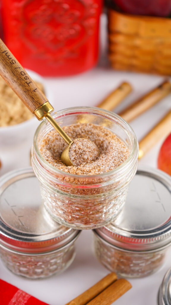 A small glass jar filled with cinnamon sugar mix, perfect for making Apple Cider Mix, with a small measuring spoon inside, surrounded by jars, cinnamon sticks, and apples in the background.