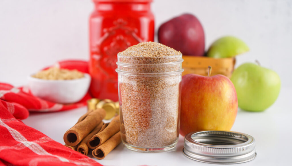 A glass jar filled with cinnamon sugar sits on a white surface near whole apples, cinnamon sticks, a red jar, a bowl of brown sugar, and a homemade Apple Cider Mix.