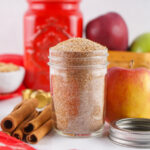 A glass jar filled with cinnamon sugar sits on a white surface near whole apples, cinnamon sticks, a red jar, a bowl of brown sugar, and a homemade Apple Cider Mix.