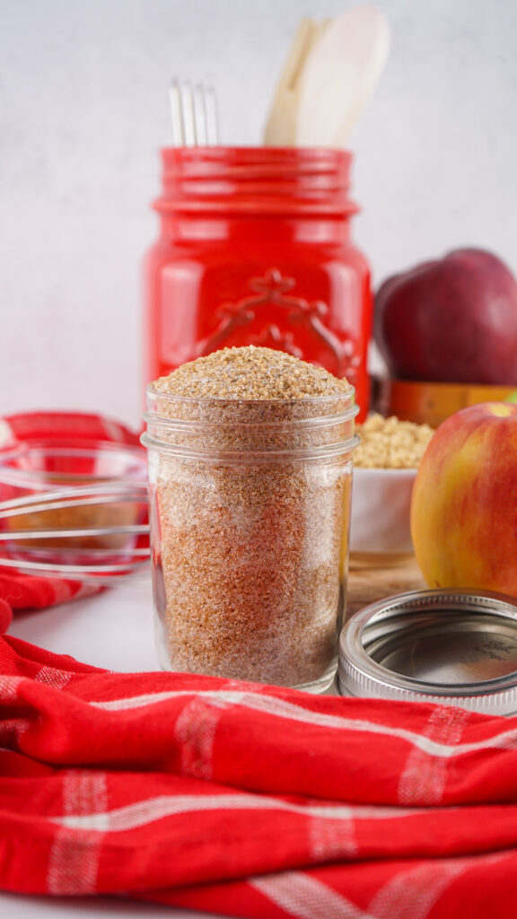 A glass jar filled with brown granulated sugar sits on a white surface, surrounded by a red cloth, apples, an Apple Cider Mix container, and kitchen utensils in the background.
