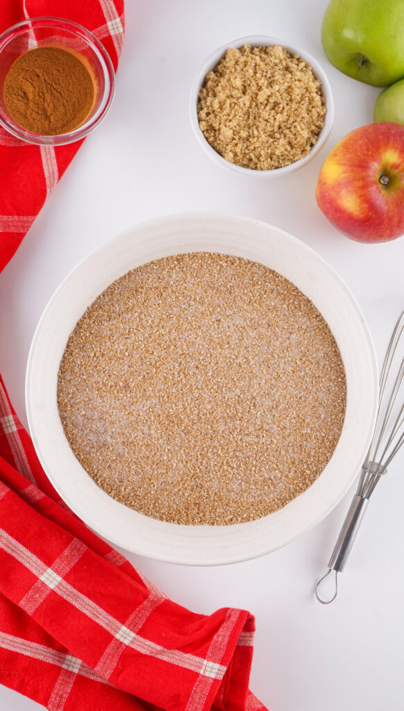 A white bowl filled with a brown sugar and spice mixture—perfect for your Apple Cider Mix—sits on a white surface beside apples, a bowl of brown sugar, cinnamon, a whisk, and a red checkered cloth.