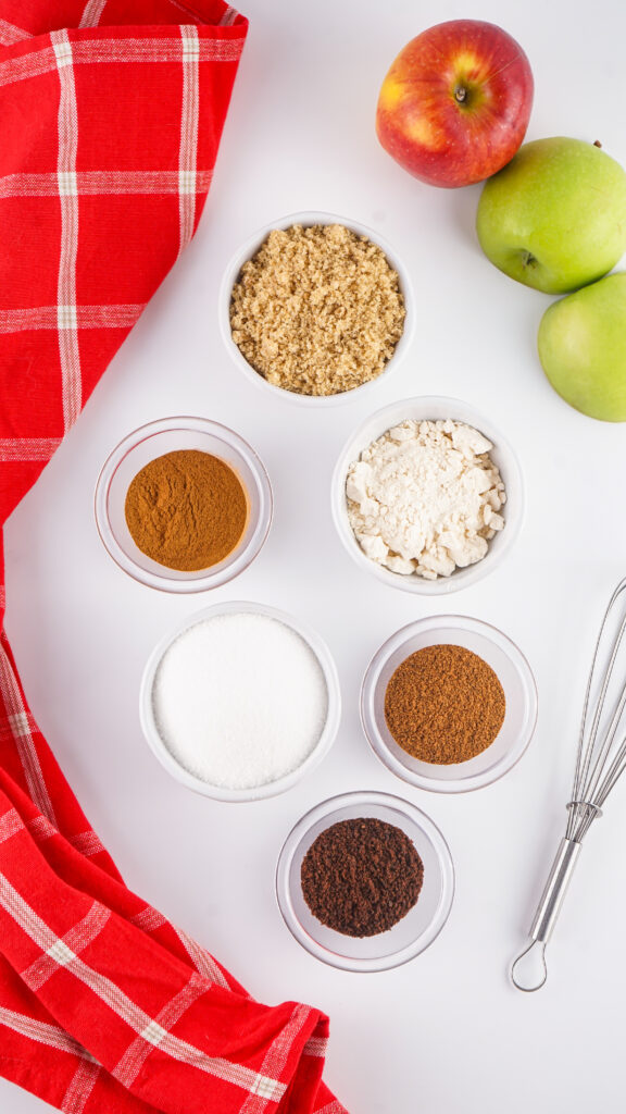 Six small bowls with brown sugar, flour, white sugar, cinnamon, nutmeg, and ground cloves—perfect for making an Apple Cider Mix—sit beside apples, a red plaid cloth, and a metal whisk on a white surface.